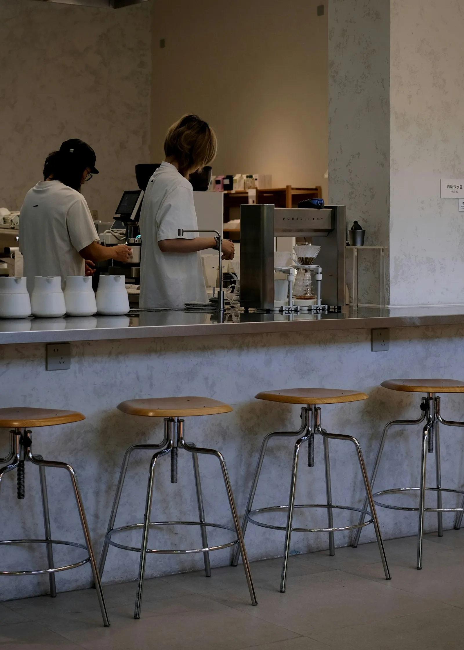 Two baristas working behind a bright café counter lined with stools